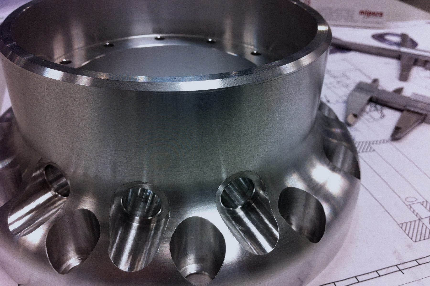 A machined metal housing resting on its engineering drawing beside a caliper